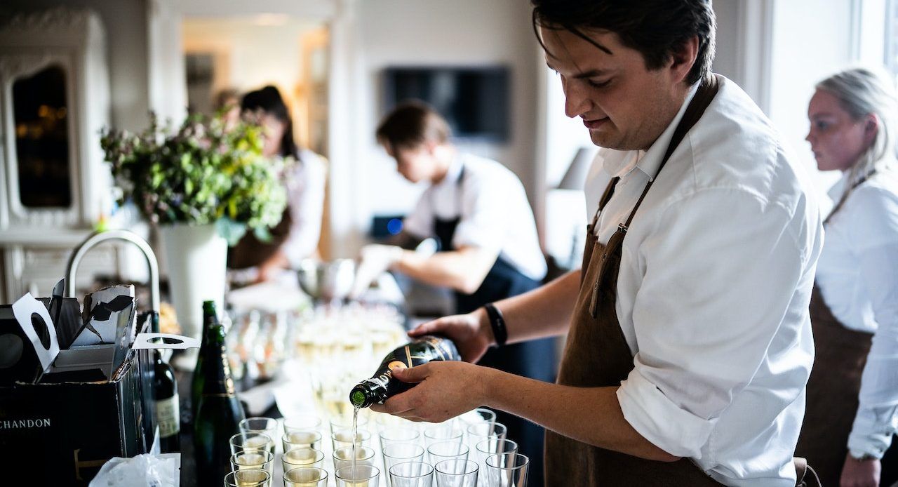 Ein Kellner schenkt Sekt in Gläser ein, während ein ganzes Kellnerteam im Hintergrund eines Restaurants zu sehen ist.